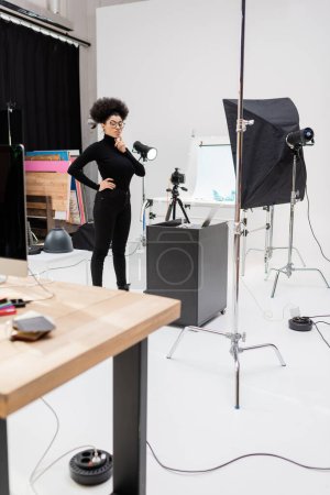 nachdenklicher afrikanisch-amerikanischer Content-Macher steht mit der Hand auf hüftnahem Laptop und Softbox-Reflektor im Fotostudio