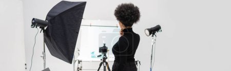 vista posterior del administrador de contenido afroamericano en ropa negra cerca de equipos de iluminación y cámara digital en el estudio de fotografía, pancarta