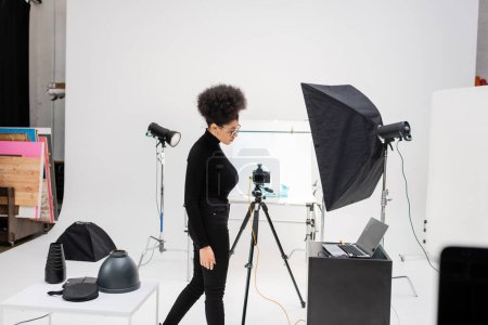 Foto de Vista lateral del productor de contenido afroamericano en ropa negra cerca de la computadora portátil y equipo de iluminación en el moderno estudio de fotografía - Imagen libre de derechos