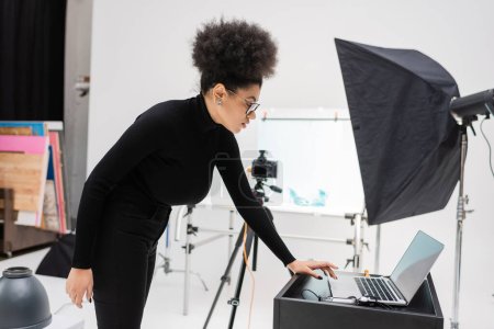 Téléchargez les photos : Vue latérale du fabricant de contenu afro-américain à l'aide d'un ordinateur portable avec écran vide près du réflecteur softbox dans le studio photo - en image libre de droit