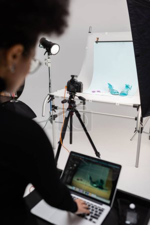 Foto de Productor de contenido afroamericano borrosa trabajando en el ordenador portátil cerca de la cámara digital y el calzado de moda en la mesa de fotos en el estudio de fotos - Imagen libre de derechos