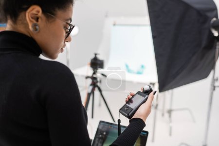 afrikanisch-amerikanischer Content-Produzent im schwarzen Rollkragen schaut auf Belichtungsmesser im verschwommenen Fotostudio