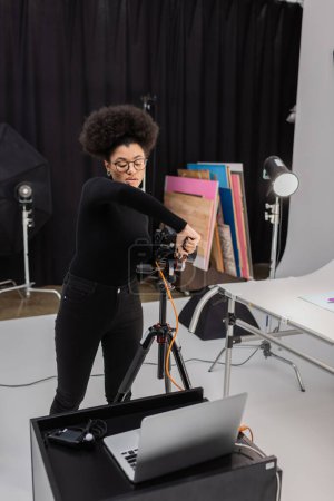 Afrikanisch-amerikanischer Content-Produzent in Brille montiert Digitalkamera auf Stativ in der Nähe von Laptop im Fotostudio