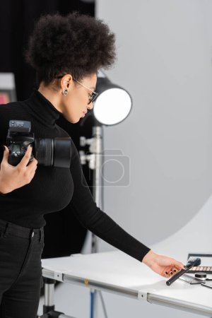 Photo for African american content producer with digital camera holding cosmetic brush near cosmetics on shooting table in photo studio - Royalty Free Image