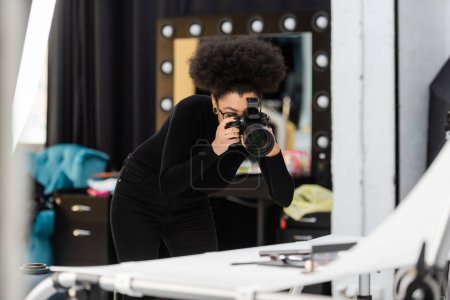 african american content maker taking photo of decorative cosmetics on blurred shooting table in photo studio
