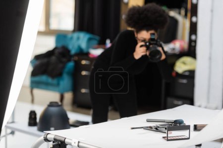 blurred african american content maker taking photo of beauty tools and decorative cosmetics on shooting table in studio
