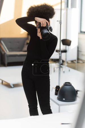 african american content producer in black clothes taking picture on professional digital camera while working in photo studio