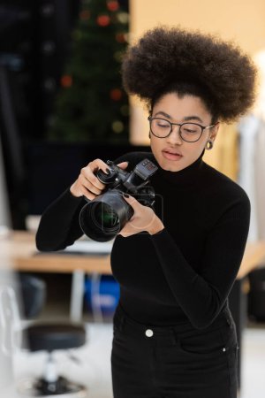 stylish african american photographer in eyeglasses and black turtleneck working with digital camera in photo studio