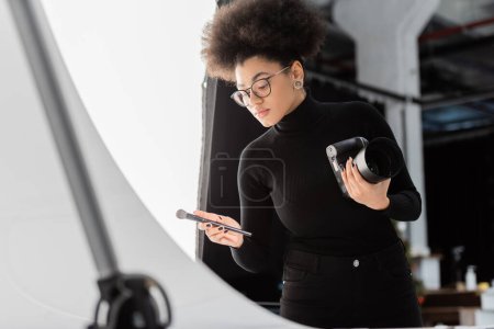 african american content manager holding digital camera and cosmetic brush in photo studio
