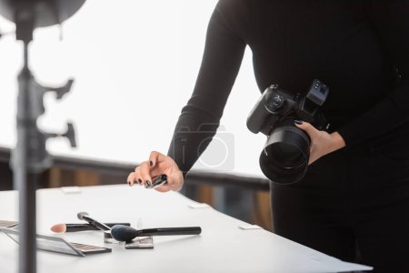 partial view of african american photographer with digital camera holding mascara near eye shadows and cosmetic brushes on shooting table in studio