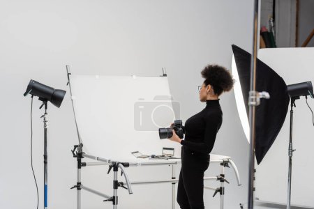 Foto de Vista lateral del fabricante de contenido afroamericano con cámara digital mirando cosméticos decorativos y herramientas de belleza en el estudio de fotografía - Imagen libre de derechos