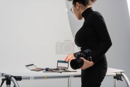 african american content maker with digital camera touching eye shadows palette near decorative cosmetics on shooting table in photo studio
