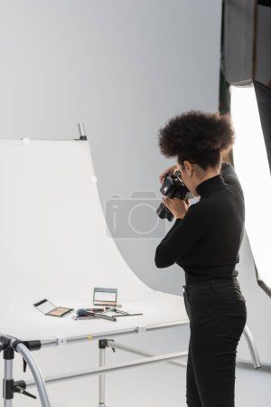 Foto de Fotógrafo afroamericano en cuello alto negro tomando foto de cosméticos decorativos en mesa de fotos en estudio fotográfico - Imagen libre de derechos
