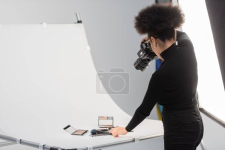 african american photographer taking photo of beauty tools and decorative cosmetics on shooting table in photo studio