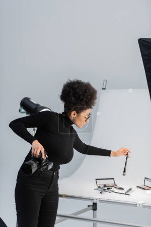 african american content producer holding digital camera and cosmetic brush near shooting table in photo studio