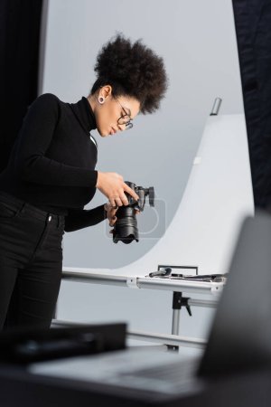 african american photographer in black turtleneck and eyeglasses looking at digital camera near decorative cosmetics in photo studio