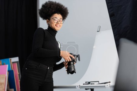 joyful african american photographer in eyeglasses smiling at camera near decorative cosmetics and beauty tools in photo studio