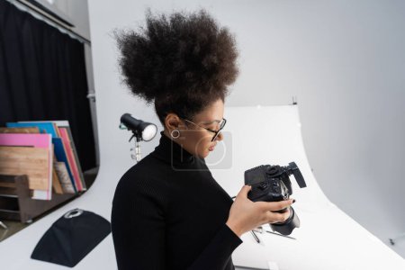 Téléchargez les photos : Vue de côté du photographe afro-américain regardant l'écran de l'appareil photo numérique dans le studio photo - en image libre de droit