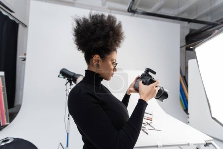 Téléchargez les photos : Vue latérale du créateur de contenu afro-américain à col roulé noir regardant un appareil photo numérique dans un studio photo - en image libre de droit