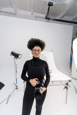 Téléchargez les photos : Joyeuse gestionnaire de contenu afro-américain avec appareil photo numérique près des projecteurs et de la table de tir dans le studio photo moderne - en image libre de droit