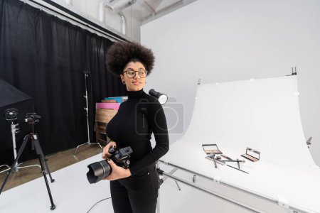 happy african american content maker with digital camera looking away near decorative cosmetics on shooting table in photo studio