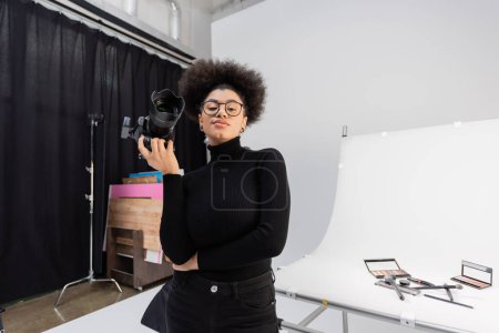 productor de contenido afroamericano sonriente sosteniendo la cámara digital mientras está de pie en la mesa de rodaje con cosméticos decorativos en el estudio de fotos
