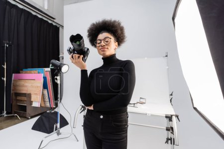 positive african american content maker in black turtleneck and eyeglasses standing with professional digital camera in photo studio