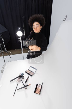fotógrafo afroamericano en gafas tomando fotos de herramientas de belleza y cosméticos en la mesa de fotos en el estudio de fotografía