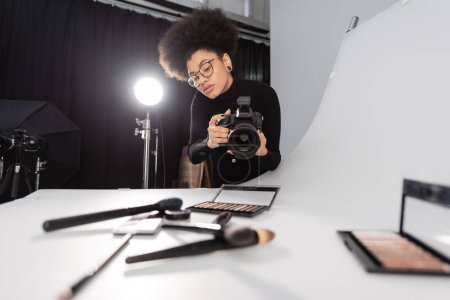 african american content manager in eyeglasses taking picture of blurred beauty tools and decorative cosmetics in photo studio