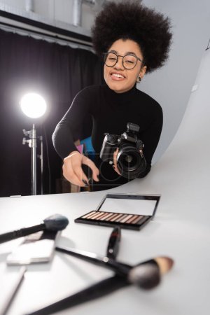 cheerful african american photographer in eyeglasses looking at camera near decorative cosmetics on shooting table in photo studio