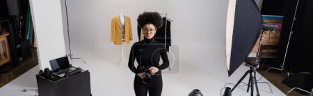 Téléchargez les photos : Afro-américain fabricant de contenu avec appareil photo numérique près de la collection de vêtements élégants et ordinateur portable dans le studio photo, bannière - en image libre de droit