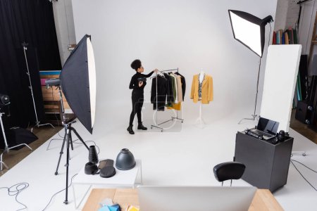 in voller Länge afrikanisch-amerikanischer Content-Produzent arbeitet mit neuer Kollektion modischer Kleidung in der Nähe von Scheinwerfern im Fotostudio