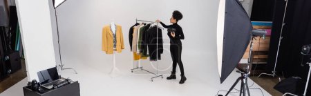 pleine longueur de fabricant de contenu afro-américain près de la nouvelle collection de vêtements élégants et projecteurs dans le studio photo, bannière