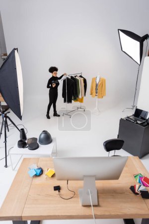 full length of african american content manager near rail rack with trendy clothing in photo studio with computers and lighting equipment