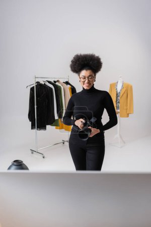 Téléchargez les photos : Joyeux producteur de contenu afro-américain avec appareil photo numérique près de la collection de vêtements à la mode dans le studio photo - en image libre de droit