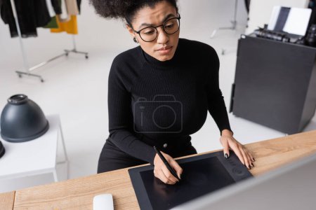 focused african american retoucher in black clothes and eyeglasses using graphic tablet while working in photo studio