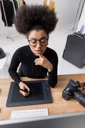high angle view of thoughtful african american retoucher in eyeglasses working on graphic tablet and looking at monitor in photo studio