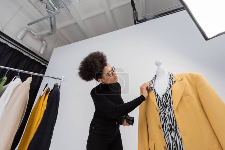 low angle view of african american photographer touching fashionable blazer on mannequin in photo studio