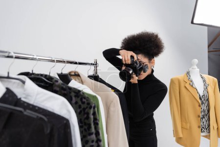 african american photographer taking picture of fashionable clothes collection on rail rack in photo studio