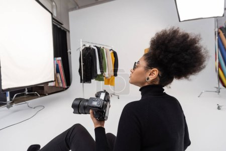 fotógrafo afroamericano con cámara digital mirando ropa de moda en estudio de fotos