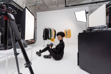 pleine longueur de joyeux photographe afro-américain regardant l'appareil photo tout en étant assis près de la collection de vêtements élégants dans le studio de photo