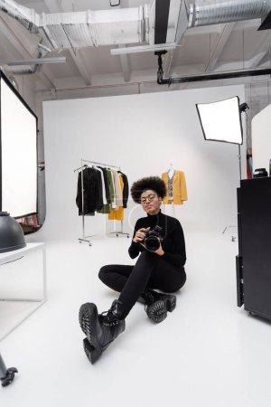 Foto de Longitud completa de fotógrafo afroamericano en ropa elegante negro sentado con cámara digital en estudio de fotografía moderna - Imagen libre de derechos