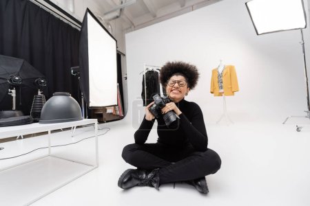 productor de contenido afroamericano lleno de alegría con cámara digital sentada con los ojos cerrados en el moderno estudio de fotografía