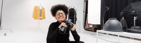 Foto de Alegre afroamericano fotógrafo en gafas sonriendo con los ojos cerrados mientras está sentado en el estudio de fotografía, pancarta - Imagen libre de derechos