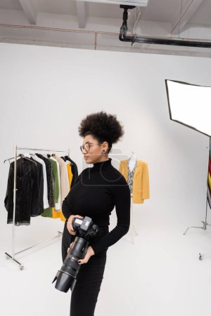Afrikanischer amerikanischer Fotograf mit Brille und schwarzem Rollkragen steht mit Digitalkamera in der Nähe einer Sammlung trendiger Kleidung im Fotostudio