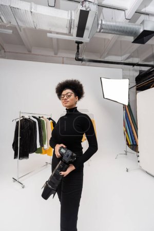 positive african american photographer in eyeglasses standing with digital camera near rail rack with trendy clothes in photo studio