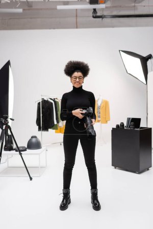 pleine longueur de joyeux fabricant de contenu afro-américain en vêtements noirs debout avec appareil photo numérique dans le studio photo moderne