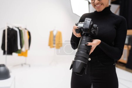 partial view of cheerful african american photographer in black turtleneck holding professional digital camera in blurred photo studio