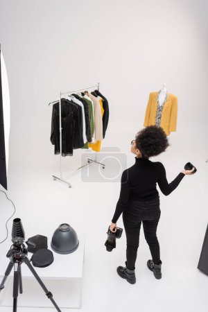 Hochwinkelaufnahme eines afrikanisch-amerikanischen Content-Herstellers mit Digitalkamera und Fotoobjektiv bei der Betrachtung einer modischen Kleidersammlung im Fotostudio