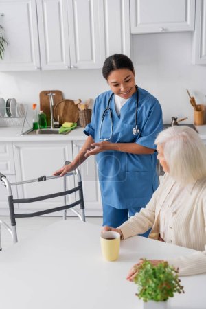 fröhliche Krankenschwester in blauer Uniform, die mit der Hand auf Rollator zeigt, während sie neben einer Seniorin mit grauen Haaren steht 